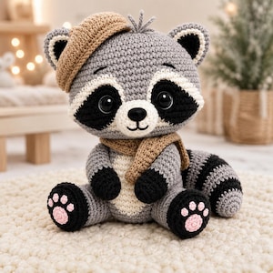 May include: A crocheted raccoon plush toy featuring grey and white yarn, wearing a tan beret and a matching tan scarf. The raccoon has black eye patches, a black nose, black paws with pink paw pads, and a striped tail. It is seated upright.