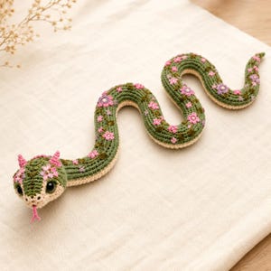Puede incluir: Un juguete de serpiente tejido a ganchillo con cuerpo verde y parte inferior de color crema. La serpiente está decorada con pequeños bordados florales rosas y morados, tiene dos pequeños cuernos rosas en la cabeza, ojos negros y lengua bífida.