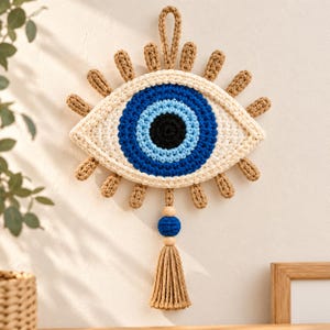 May include: A crocheted wall hanging shaped like an eye with concentric circles in black, bright blue, and cream yarn. The design features radiating tan yarn lashes, a top hanging loop, and a dangling tan tassel with a blue bead and two wooden beads.