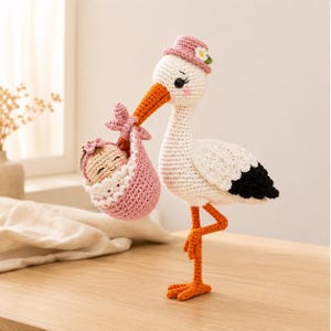 Puede incluir: Una figura de cigüeña tejida a ganchillo a mano con plumaje blanco, puntas de alas negras y pico naranja. La cigüeña lleva un pequeño sombrero rosa con una flor blanca y carga un bulto tejido en rosa que contiene una pequeña muñeca.