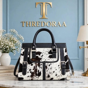 May include: A structured handbag featuring a black and white cowhide print pattern, black leather trim, and two vertical straps with silver-toned buckles on the front. The bag has a top handle and a detachable shoulder strap, with visible stitching.