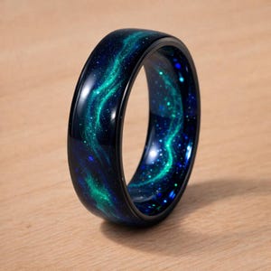 May include: A polished ring featuring a deep navy blue and black base with a swirling, luminous green nebula pattern that mimics the appearance of a galaxy. The surface is smooth and glossy, displaying scattered bright specks of light throughout.