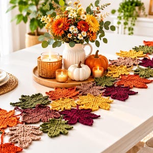 May include: A table runner composed of interconnected crocheted maple and oak leaves in autumn colors including orange, yellow, dark red, olive green, and brown. The runner is displayed on a white table alongside a floral centerpiece and pumpkins.