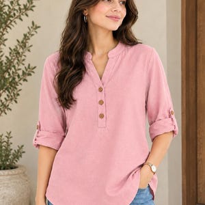 May include: A dusty pink long-sleeve shirt featuring a mandarin collar, a V-neckline with three brown buttons, and roll-tab sleeves. The shirt has a curved hemline and is paired with blue denim jeans, creating a casual look with a soft texture.