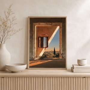 May include: A framed wall art print depicting an open doorway looking out onto a landscape at sunset. The scene features a stone archway, an open window with dark shutters, a terracotta floor, and a distant view of hills and a single cypress tree.