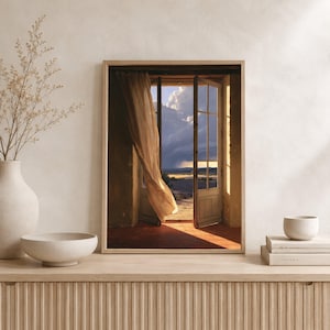 May include: A framed wall art print depicting an open doorway with a sheer beige curtain blowing in the breeze, revealing a landscape with a dramatic cloudy sky and golden light, displayed in a room with a vase, bowl, and stacked books.