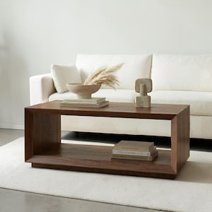 May include: A rectangular dark brown wooden coffee table with an open shelf design. The table features a smooth finish and clean lines. On the top surface sits a beige bowl with dried grasses and a small abstract sculpture, plus two stacked books.