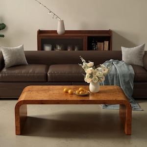 May include: A low-profile, rectangular coffee table with a light brown burl wood veneer finish and curved waterfall edges. The table surface holds a white ceramic vase filled with white flowers and a small bowl containing yellow citrus fruits.
