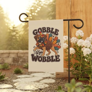 Op de afbeelding: Een crèmekleurige tuin vlag met een bruine mopshond in een kalkoenkostuum met een kleurrijke staart met patronen. De tekst GOBBLE til you WOBBLE staat in donkerbruine letters boven en onder de illustratie van de hond.
