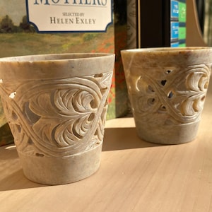 May include: Two beige stone candle holders featuring intricate, hand-carved openwork leaf patterns. Each cylindrical vessel has a smooth, solid base and a wide, open top, showcasing the natural, mottled texture of the carved stone material.
