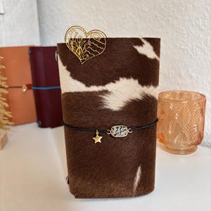May include: A brown and white cowhide leather notebook cover featuring a gold-toned metal heart-shaped bookmark with a floral cutout design. A black elastic band wraps around the cover, holding a small gold star charm and a rectangular stone bead.