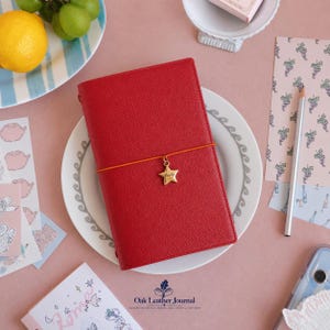 May include: A red leather journal with a textured surface, held closed by an orange elastic band featuring a gold-colored star charm. The journal is centered on a white plate with a gray decorative border, surrounded by stationery items.