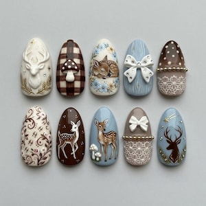 May include: A set of ten almond-shaped press-on nails featuring intricate designs including a white deer head, brown plaid with a mushroom, a sleeping fawn, a white bow, lace patterns, floral swirls, standing deer, and small rhinestones.