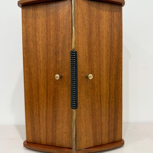 May include: A wooden cabinet with two doors featuring a vertical brass handle with a black ribbed grip. The cabinet has a curved top and base, with two small brass knobs on the doors. The wood grain is visible across the front panels.