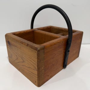 Könnte beinhalten: Ein rechteckiger Holzkasten mit zwei offenen Fächern und einem stabilen, bogenförmigen schwarzen Metallgriff, der mit sichtbaren Schrauben an den Seiten befestigt ist. Das Holz zeigt eine natürliche Maserung mit einem warmen, mittelbraunen Finish.