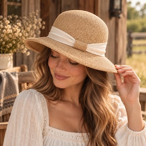 Puede incluir: Un sombrero de paja color canela con una copa redondeada y un ala de ancho medio. El sombrero presenta una banda de tela ancha de color blanco roto envuelta alrededor de la base de la copa, atada con un pequeño detalle de nudo central.