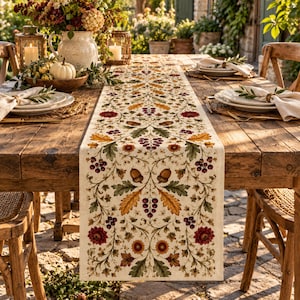 May include: A cream-colored table runner featuring a repeating botanical pattern of red flowers, orange and green oak leaves, brown acorns, and clusters of dark red berries, centered on a rustic wooden dining table with place settings.