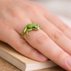 May include: A ring shaped like a small green frog with gold-toned legs and feet. The frog is positioned so its body wraps around the finger, with its front legs extending forward and its back legs resting along the side of the band.