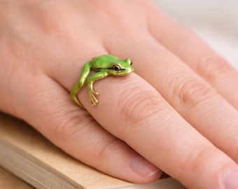 May include: A ring shaped like a small green frog with gold-toned legs and feet. The frog is positioned so its body wraps around the finger, with its front legs extending forward and its back legs resting along the side of the band.