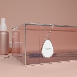 Puede incluir: Un collar con colgante en forma de lágrima plateada que presenta un inserto central blanco con el texto impreso "Emmy & Anna 09.22.2025". El colgante cuelga de una delicada cadena plateada, exhibido frente a una caja de cristal rosado.