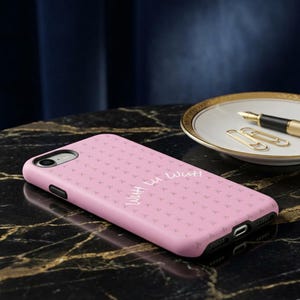 Peut inclure: Une coque de smartphone rose clair avec un motif répétitif de petits symboles sombres et le texte cursif blanc "With u wutt" imprimé au dos. La coque présente une découpe pour l'objectif de l'appareil photo et des bords noirs avec des ouvertures.
