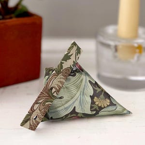 May include: A triangular paper gift box featuring a botanical pattern with shades of sage green, dark brown, and cream. The box has a folded top closure with a small circular hole, displaying intricate floral and leaf motifs across its surface.