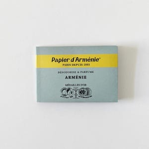 Può includere: Un libretto rettangolare di carta d'incenso Papier d'Arménie con una copertina azzurra e una striscia orizzontale gialla. Il testo riporta Papier d'Arménie, Paris Depuis 1885, Désodorise & Parfume, Arménie, Médailles d'Or, con due illustrazioni di monete circolari.