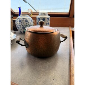 May include: A round, hammered copper pot with a matching lid and two side handles featuring twisted metal and light-colored accents. The pot has a warm, metallic finish and a rounded body, showcasing a textured surface throughout its exterior.