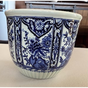 May include: A ceramic planter featuring a cream-coloured base with a repeating dark blue floral pattern arranged in vertical panels. The design includes intricate botanical motifs and a scalloped border near the base, with a wide, smooth rim.