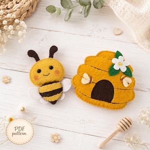May include: A felt bee plush with black antennae, black eyes, and pink cheeks, alongside a yellow felt beehive featuring a white flower with green leaves and two small bees. A wooden honey dipper is positioned near the felt items.