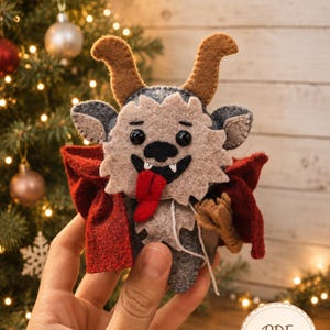Op de afbeelding: Een handgemaakt vilt ornament dat een wezen voorstelt met bruine hoorns, een beige gezicht, zwarte ogen en een brede grijns met een uitstekende rode tong. Het figuurtje draagt een donkerrode cape en heeft grijze vilten details.