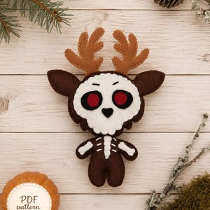 May include: A felt plush toy shaped like a deer with a skull-like face, red eyes, and brown antlers. The body is dark brown with a white ribcage design stitched on the front. A small orange pumpkin-shaped label reads PDF pattern.