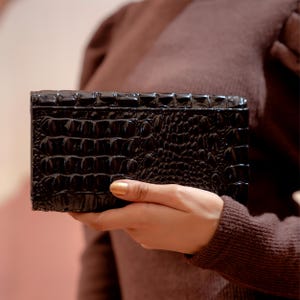 Puede incluir: Una cartera rectangular con un acabado brillante de color marrón oscuro y un patrón texturizado de piel de cocodrilo. La cartera presenta un diseño estructurado con cierre de solapa, mostrando una superficie escamosa distintiva.