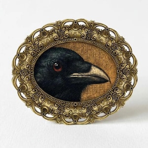 May include: An oval-shaped, antique-style frame with a detailed gold finish. Inside, a detailed painting of a black crow's head with a red eye and a light-colored beak is displayed against a wood-grain background. The frame is set against a white background.