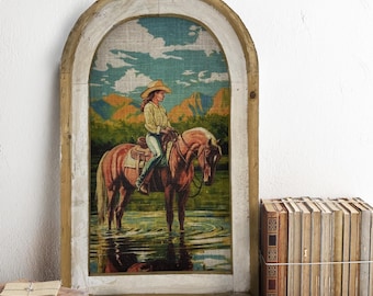 Può includere: Un dipinto incorniciato di una ragazza che cavalca un cavallo attraverso un fiume. Il dipinto è in una cornice di legno rustica con una parte superiore ad arco. Il dipinto è su uno scaffale di legno con una pila di libri e un orologio.