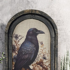 May include: A framed canvas print with a black raven perched on a branch of flowers. The raven has blue feathers and is surrounded by pink, red, and white flowers. The frame is dark brown wood with a distressed finish.