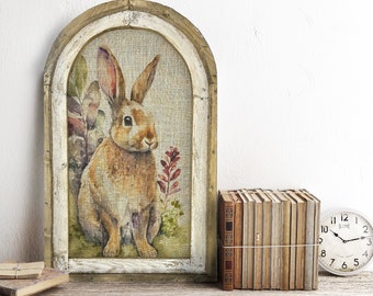Könnte beinhalten: Ein gerahmtes Gemälde eines braunen Kaninchens, das auf einer grünen Wiese mit Blumen sitzt. Der Rahmen ist aus Holz und hat eine weiße, abgenutzte Oberfläche. Das Gemälde befindet sich auf einem Holzregal mit einem Stapel Bücher und einer Uhr.