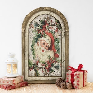 May include: A framed vintage Christmas print featuring Santa Claus with a red hat and white beard, surrounded by holly and greenery. The frame is made of wood and has a distressed white finish.