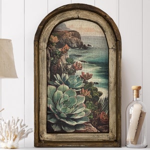 May include: A framed print of a coastal scene with succulents and a view of the ocean. The frame is brown with a whitewashed finish and has an arched top.