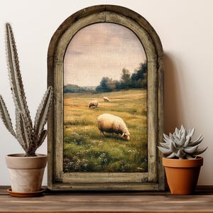 May include: A rustic wooden table displays a framed landscape painting of sheep grazing in a field. A tall cactus in a beige pot and a succulent in a terracotta pot flank the painting. The painting is framed in a distressed, arched wooden frame.