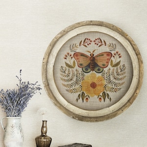 May include: A round wooden frame with a beige linen background featuring a hand-painted butterfly with orange and brown wings, a yellow flower, and green foliage.