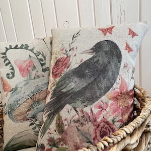 May include: Two decorative throw pillows with floral and bird designs. One pillow features a large black crow surrounded by pink and red flowers and butterflies. The other pillow has a mushroom and butterfly design. The pillows are in a woven basket.