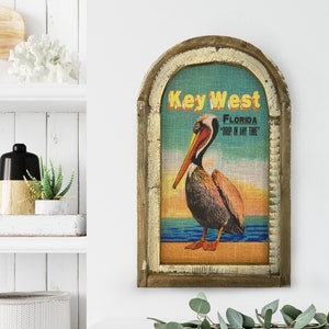 May include: Wooden framed wall art featuring a pelican illustration with the text "Key West Florida". The artwork has a distressed, vintage aesthetic with a light blue, orange, and brown color palette. The frame has a rustic, weathered appearance.