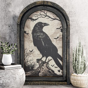 May include: A black raven silhouette on a beige canvas with a dark wood frame. The raven is perched on a tree branch with a dark background. The canvas is surrounded by a dark wood frame with a distressed finish. The image is displayed on a gray wall with two potted plants.