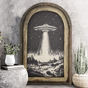 May include: A framed wall art print with a black and white illustration of a UFO flying over a desert landscape. The frame is made of wood and has a distressed finish.