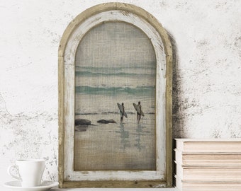 May include: A framed print of a beach scene with two surfers walking on the shore with their surfboards. The frame is a light brown wood with a white distressed finish. The print is on a canvas-like material.