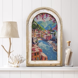 May include: A whitewashed wooden frame with an arched top, featuring a colorful print of a coastal town with a blue bay and green hills. The print is on a textured canvas.