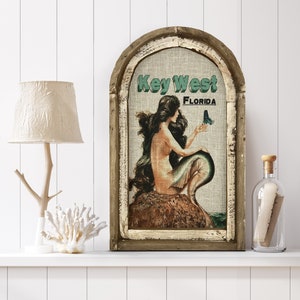 May include: A rustic wooden framed canvas print with a vintage illustration of a mermaid holding a butterfly. The canvas features the text "Key West Florida" in a teal green font.