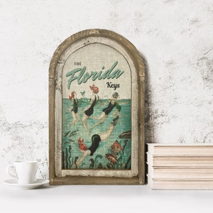 May include: A vintage-style wooden sign with a linen canvas insert featuring a retro illustration of women swimming in the ocean. The text "THE Florida Keys" is printed in green and black on the sign.
