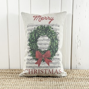 May include: A decorative throw pillow with a white background and a green holly wreath with a red bow. The pillow has the words "Merry Christmas" printed in red.